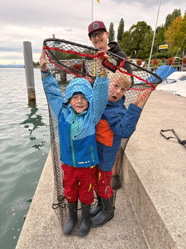 Zwei Kinder lachen im grossen Kescher, dahinter der Instruktor am See